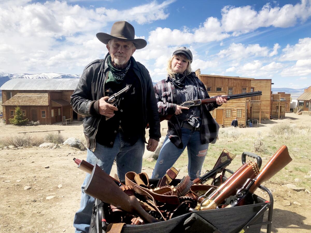 An older man and a young woman stand outdoors with an arsenal of guns, each holding a gun