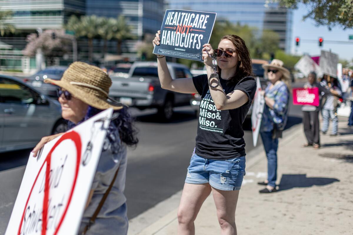 A line of protesters holding up signs, including one reading "Healthcare not handcuffs," for cars passing on a busy street.