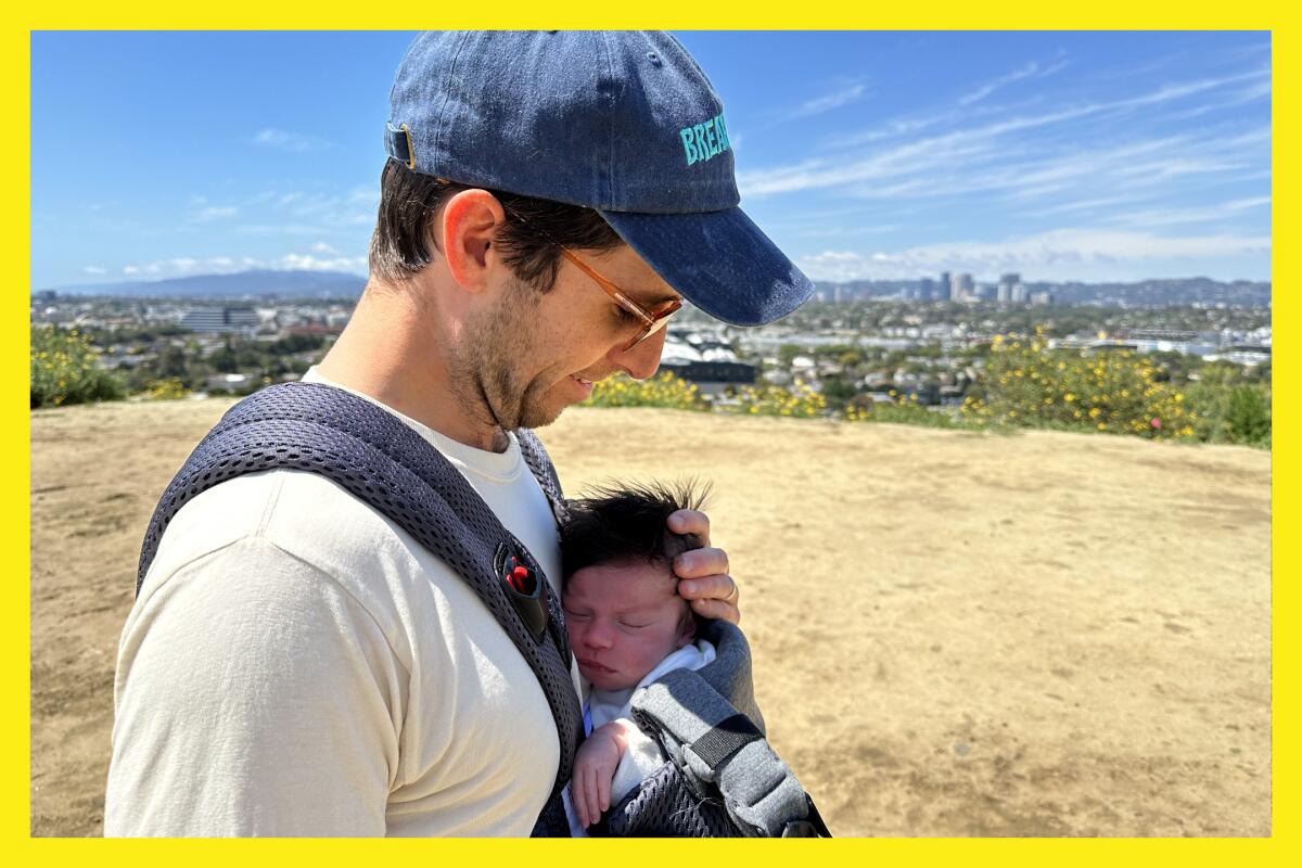 A man in a baseball cap and sunglasses carries his newborn in a BabyBjörn while hiking