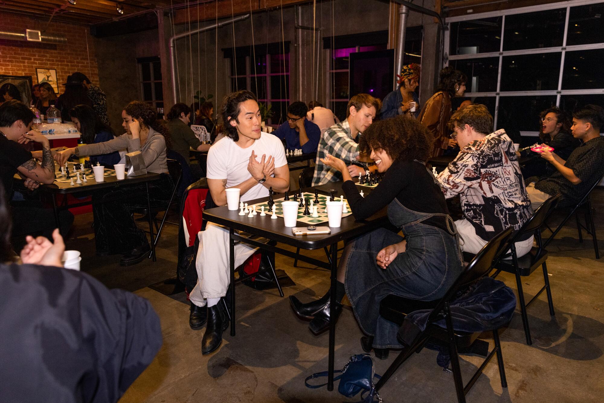 Two people in the center of a crowded room, playing chess and laughing.