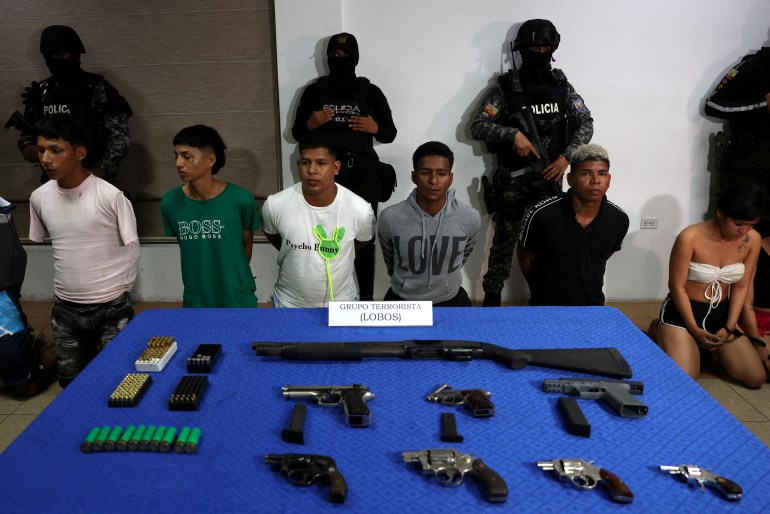 Suspects kneel in front of armed police officers. In front of them is a blue table with guns arrayed as evidence.