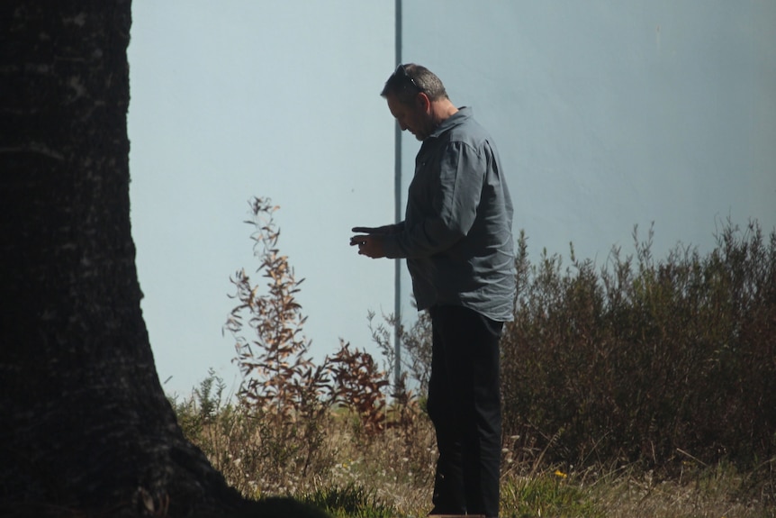 A middle-aged man stands near a tree, looking at his phone.