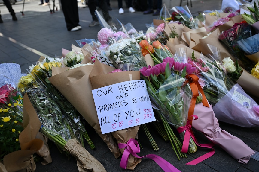 A pile of flowers on the ground. A note reads "Our hearts and prayers are with all of you."