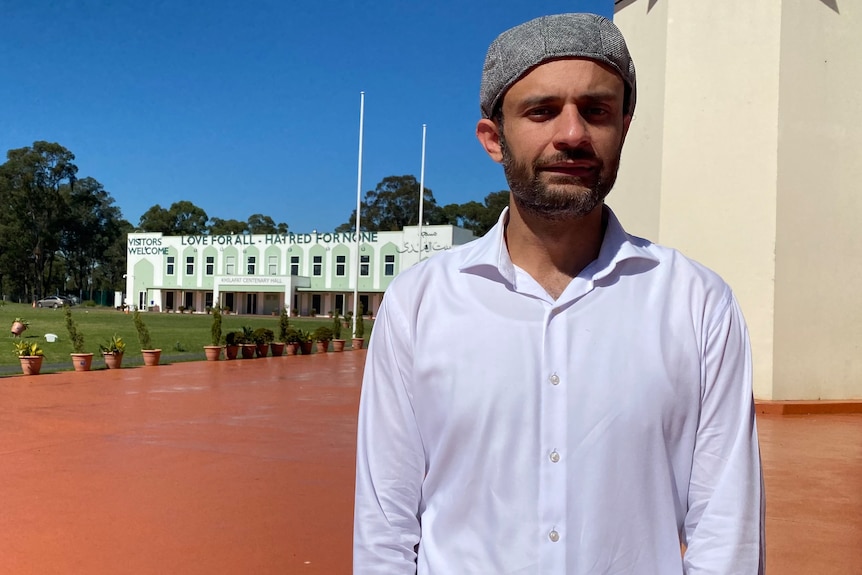 A man with a beard and wearing a hat stands outsode a mosque.