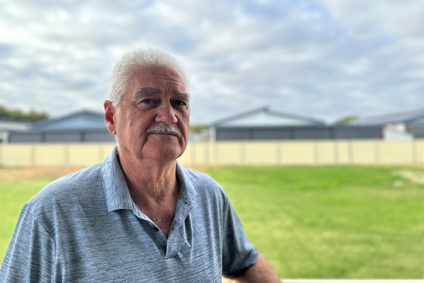 A man stands in the foreground, behind him is a green lawn and a fence.