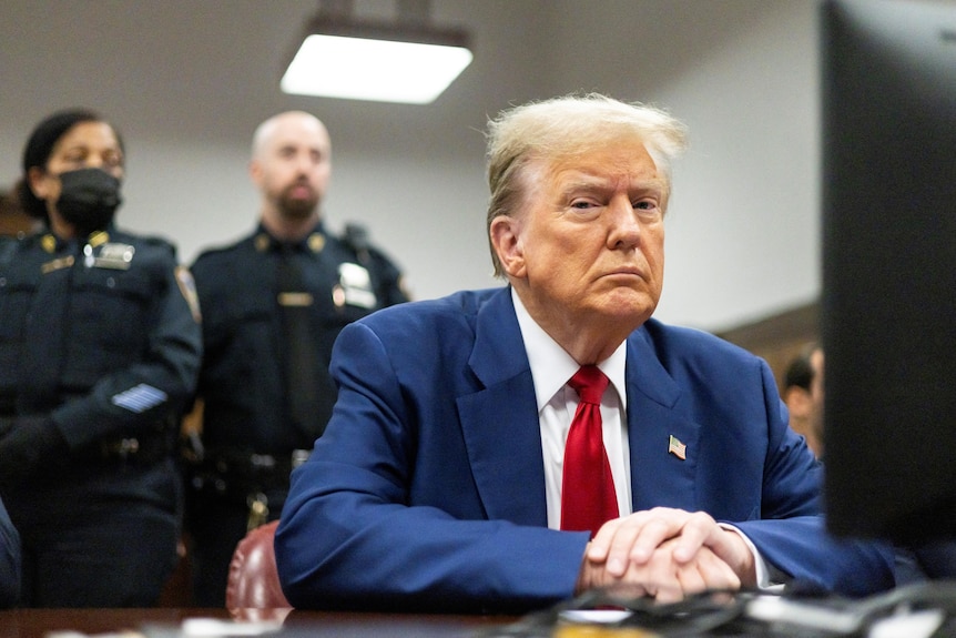 Donald Trump sits behind a desk in a blue suit and red tie looking sternly at the camera