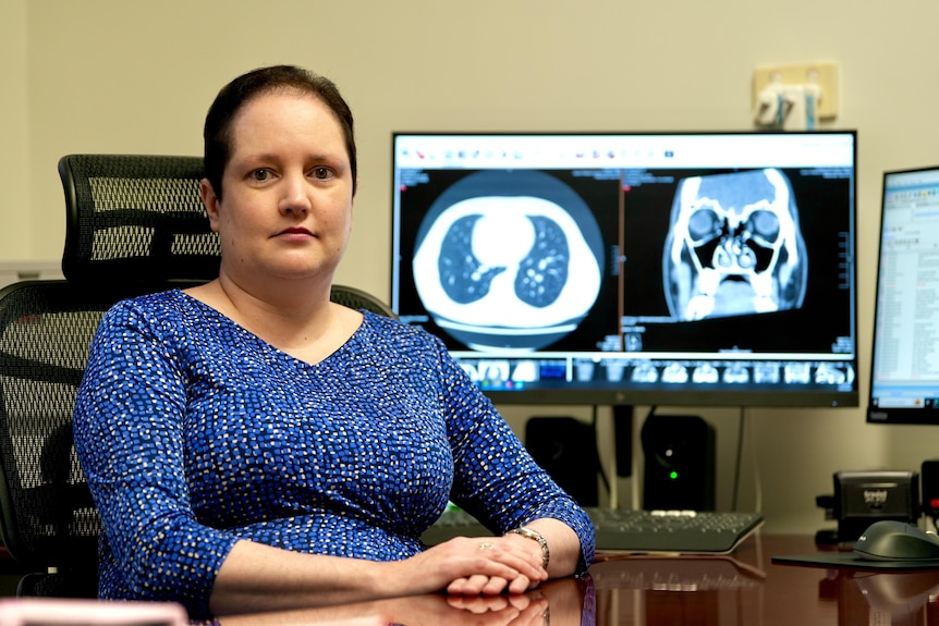 A woman with short dark hair sitting at a desk with x-ray scans on a computer screen behind her. 