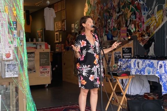 A dark-haired woman in a floral-print dress stands speaking into microphone in a small venue.