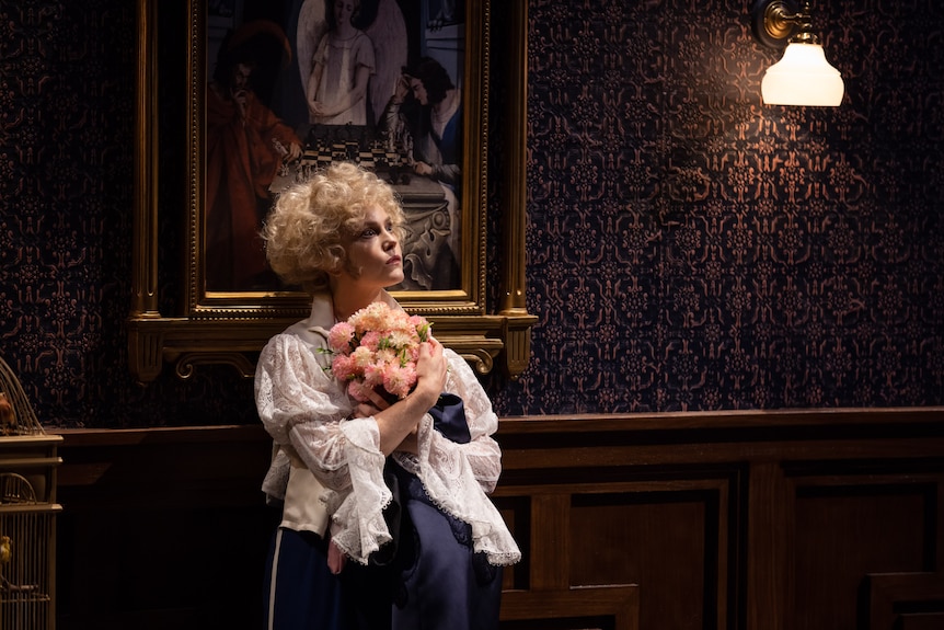 Blonde white woman dressed as man in white blouse clutches bouquet of pink flowers against lavish wallpapered interior.