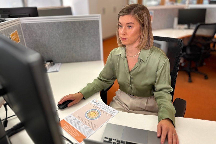 A woman with a blonde bob sits at a desk looking serious.