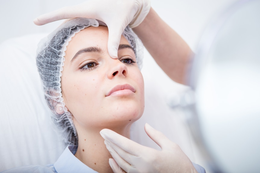 Young woman wearing hair net, while a person with white, plastic gloves surveys her face.