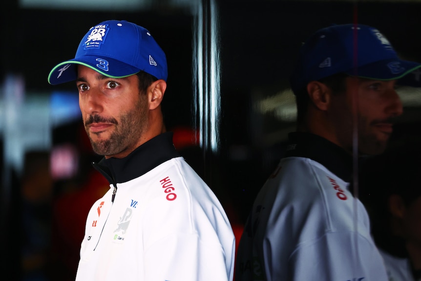 An F1 driver standing in the garage, wearing a cap, looking on the pit lane