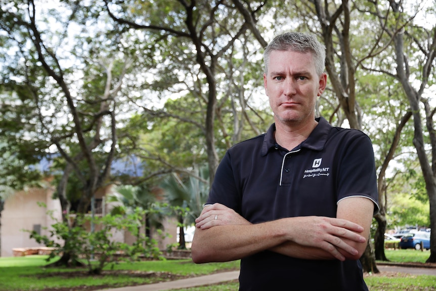 A man standing in a shady park, folding his arms and looking serious.