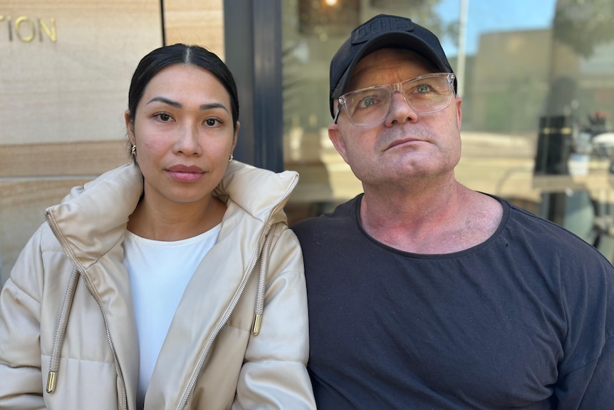 A woman and man sit next to each other. She's wearing a beige jacket and he's wearing a black shirt and cap.