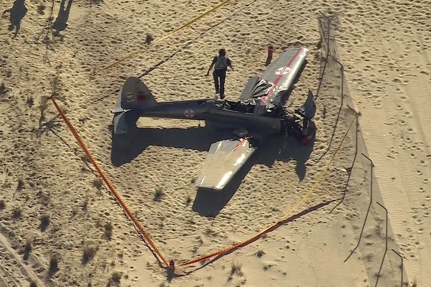 A crashed plane with a damaged nose is on sand and taped off as an emergency services work nearby.