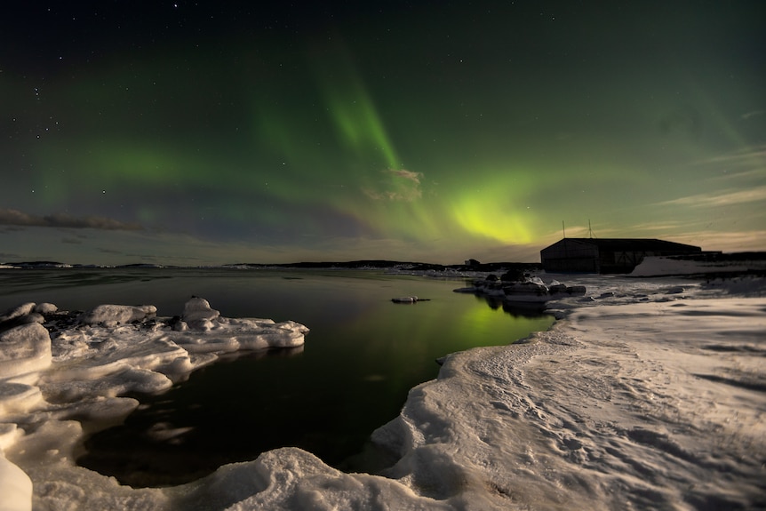 A green and yellow aurora stretches over the sky above a shed in Antarctica. 