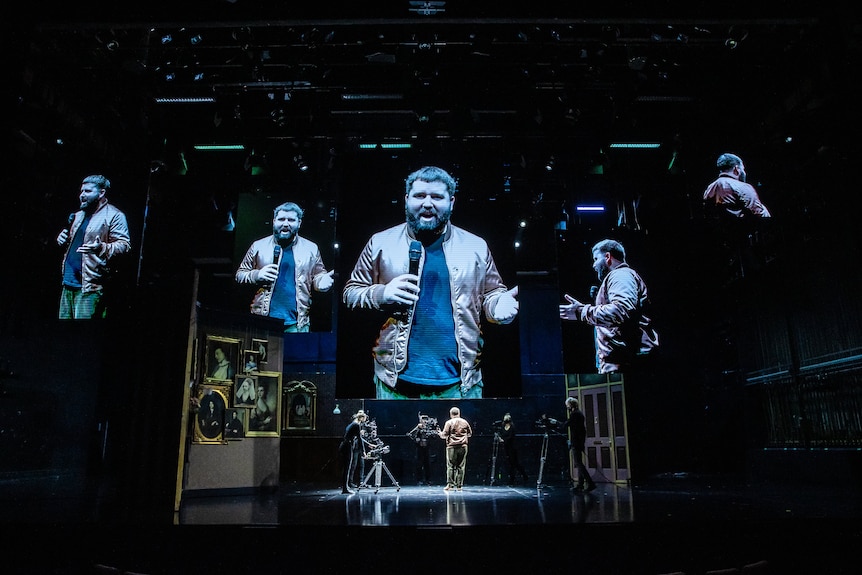 Artistic Director Kip Williams on a stage with 5 large digital screens above his head, holding a microphone.