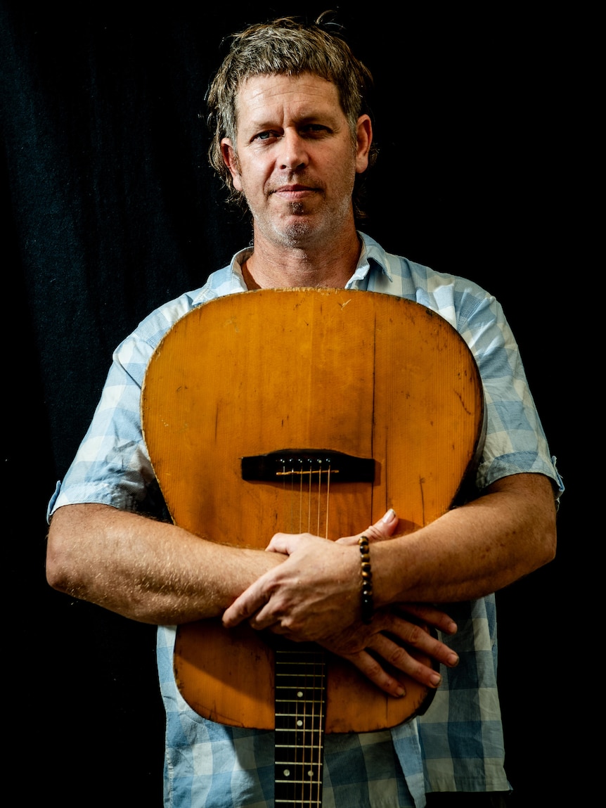 A portrait of a man wearing a light blue and white checked shirt holding his guitar upside down, close to his chest.