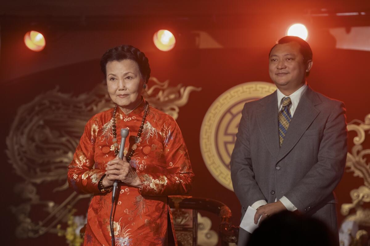 A woman in a red dress holds a microphone on a stage. A man stands behind her.