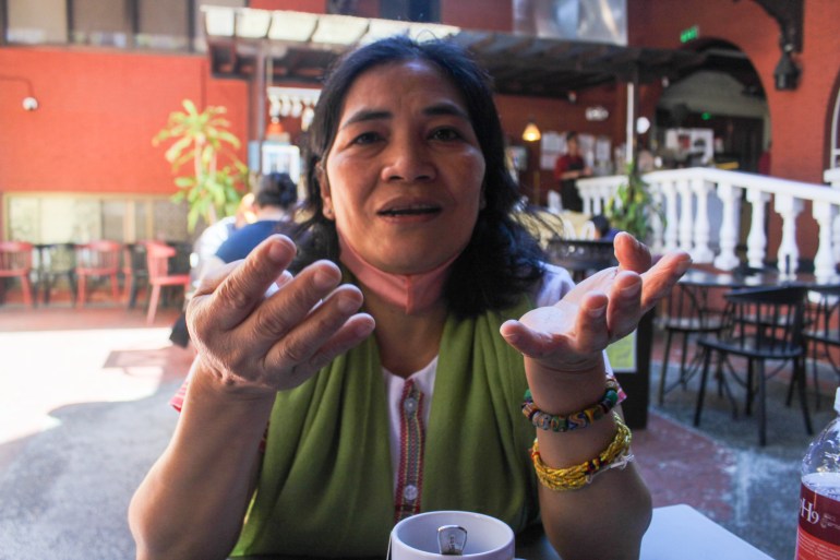 Jennifer Awingan-Taggaoa. She is sitting at a table and has a cup i front of her. She has a green scarf around her shoulders and has her arms in front of her as she speaks