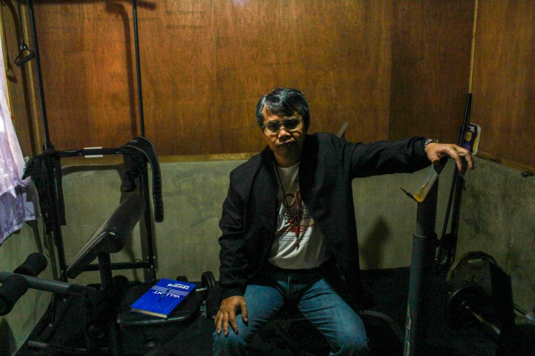 Windel in the gym. He's sitting on an exercise bench resting his hand on another piece of equipment. The walls are wooden. There's a window but it's very dark.