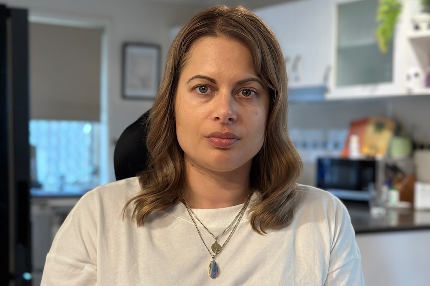 A woman sits in a kitchen, looking at the camera with a neutral expression.