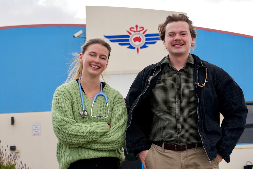 A young female doctor wearing a green sweater and a stethoscope stands next to her male counterpart