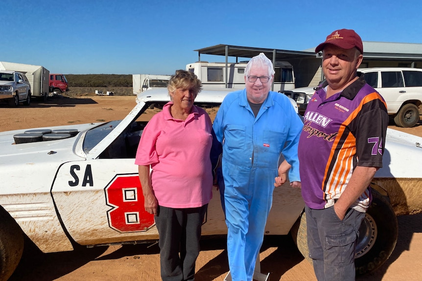 a woman and man stand beside a plastic cut out of the woman's partner in front of a white race car on a clear, blue sky day