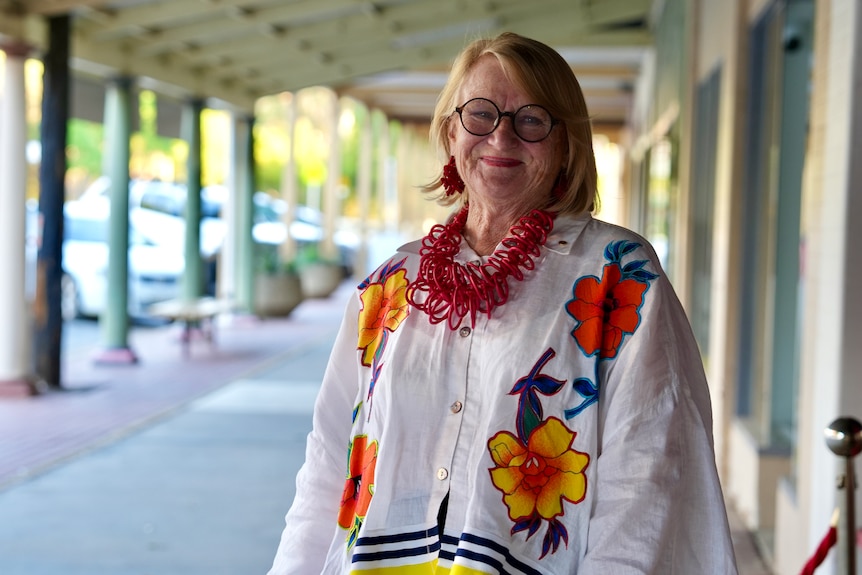 An older white woamn with blonde hair and glasses smiling at the camera on the street. 