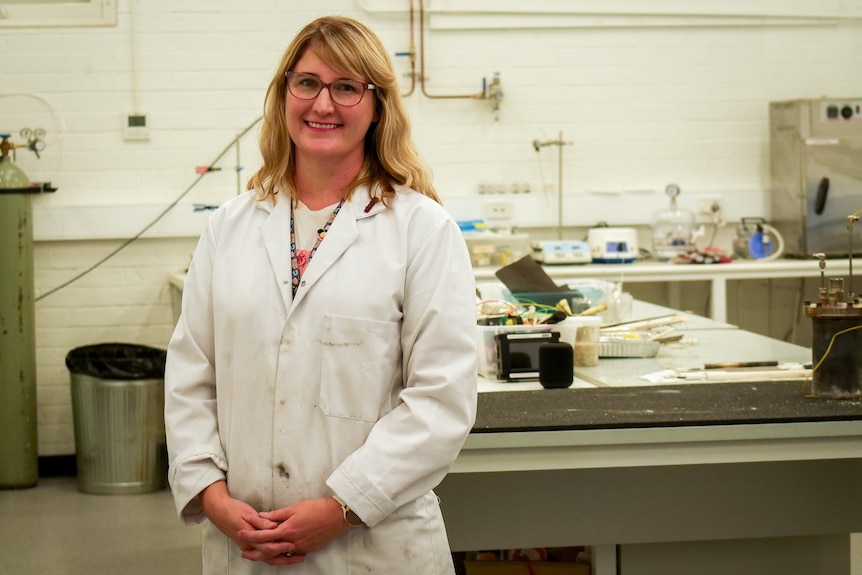 A blonde-haired woman in a white lab coats stands smiling to camera in a scientific lab.