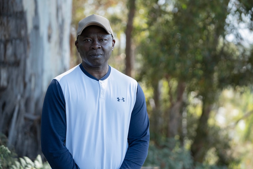 A man with a hat on, standing in front of some trees, looking at the camera.