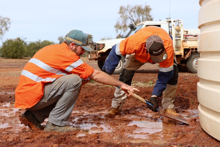 Two men in hi-vis shoveling mud near a water tank.