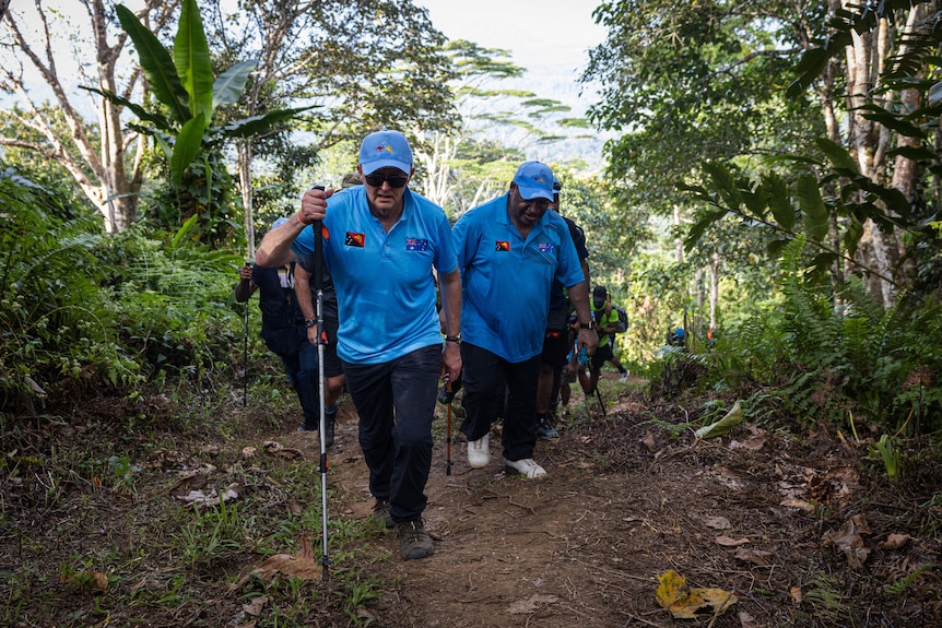 Two men in matching outfits walk closely together along a dirt track, jungle visible on either side.