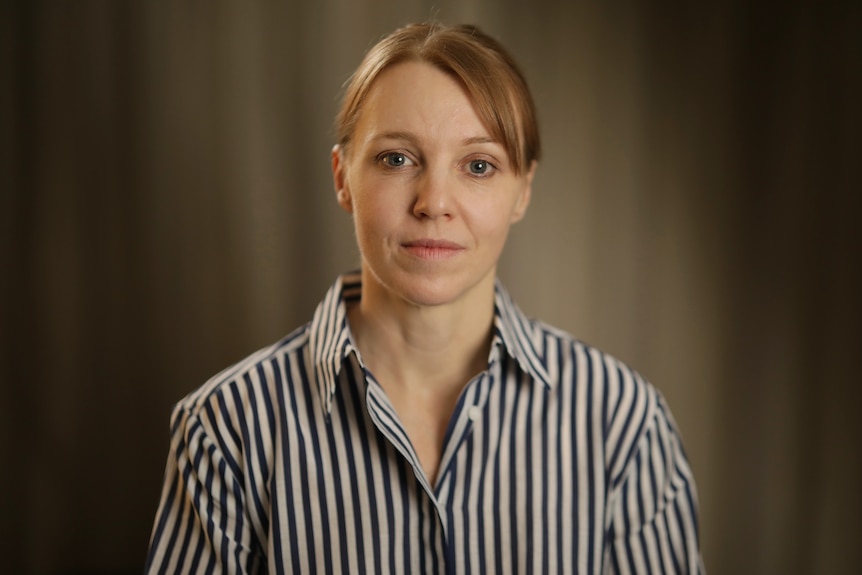 A portrait of a woman looking at the camera with a neutral expression. Behind her is plain background.