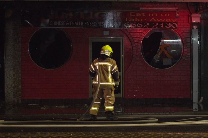 A firefighter stands with their back to the camera in front of a Chinese restaurant, pointing a fire hose at it.