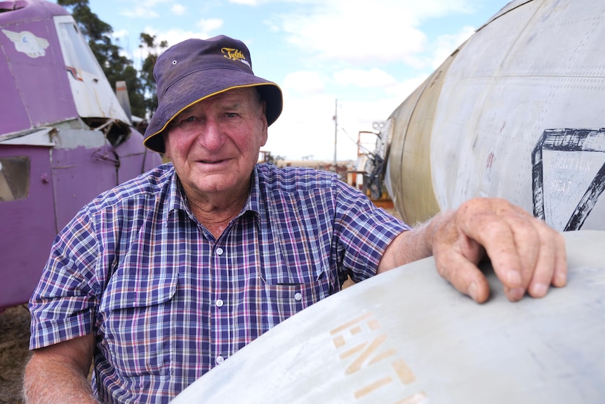 A man looks at the camera with his hand on a plane door in a plait shirt and hat against blue sky 