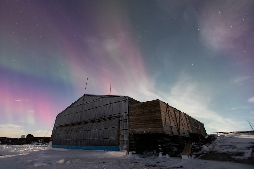 A pink and green aurora stretches over the sky above a shed in Antarctica. 