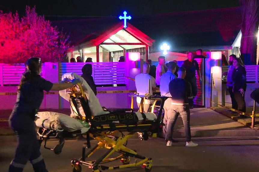 A paramedic rolls a stretcher towards a church while a crowd stand outside.