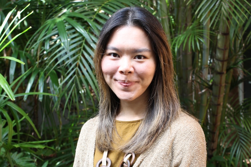 A woman with brown hari smiles against a backdrop of green plants. 