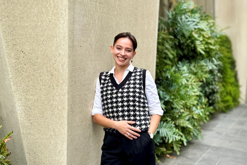 Dharani smiles in front of a sandstone wall with plants in the background. They have short brown hair and wear black and white.