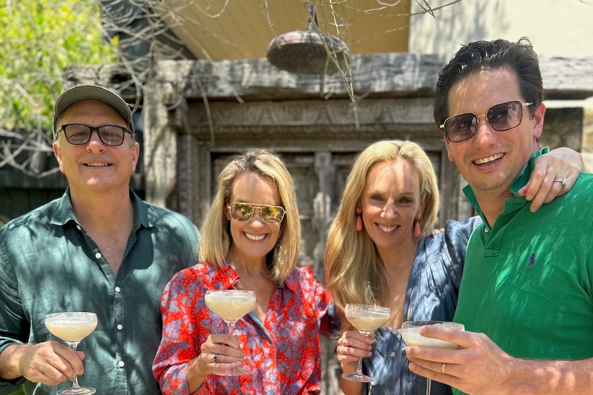 Two couples are standing together in the dappled sunlight holding cocktails and smiling.