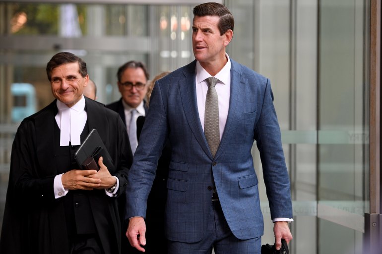 a tall man in a suit stands near a person wearing court robes