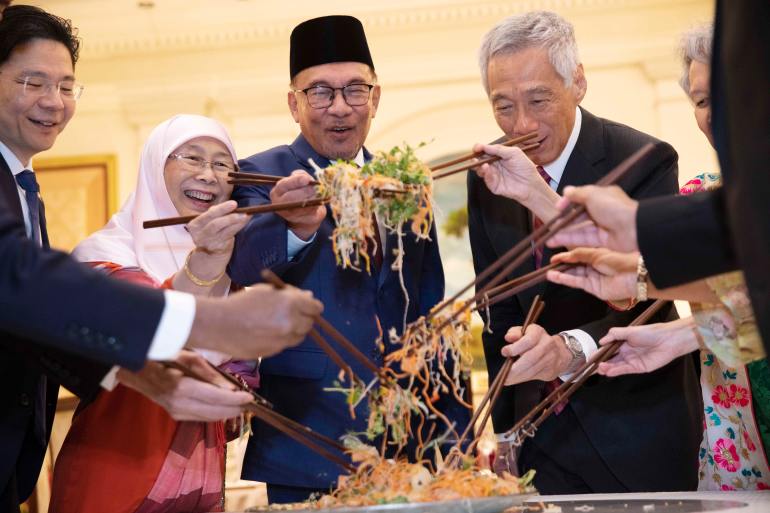 Lawrence Wong tossing 'yee sang' with Wan Azizah Wan Ismail, wife of Malaysian Prime Minister Anwar Ibrahim. Anwar Ibrahim, Singapore Prime Minister Lee Hsien Loong and his wife Ho Ching. They are around a table. The plate of yee sang is in the middie and they are using chopticks to mix the dish. 