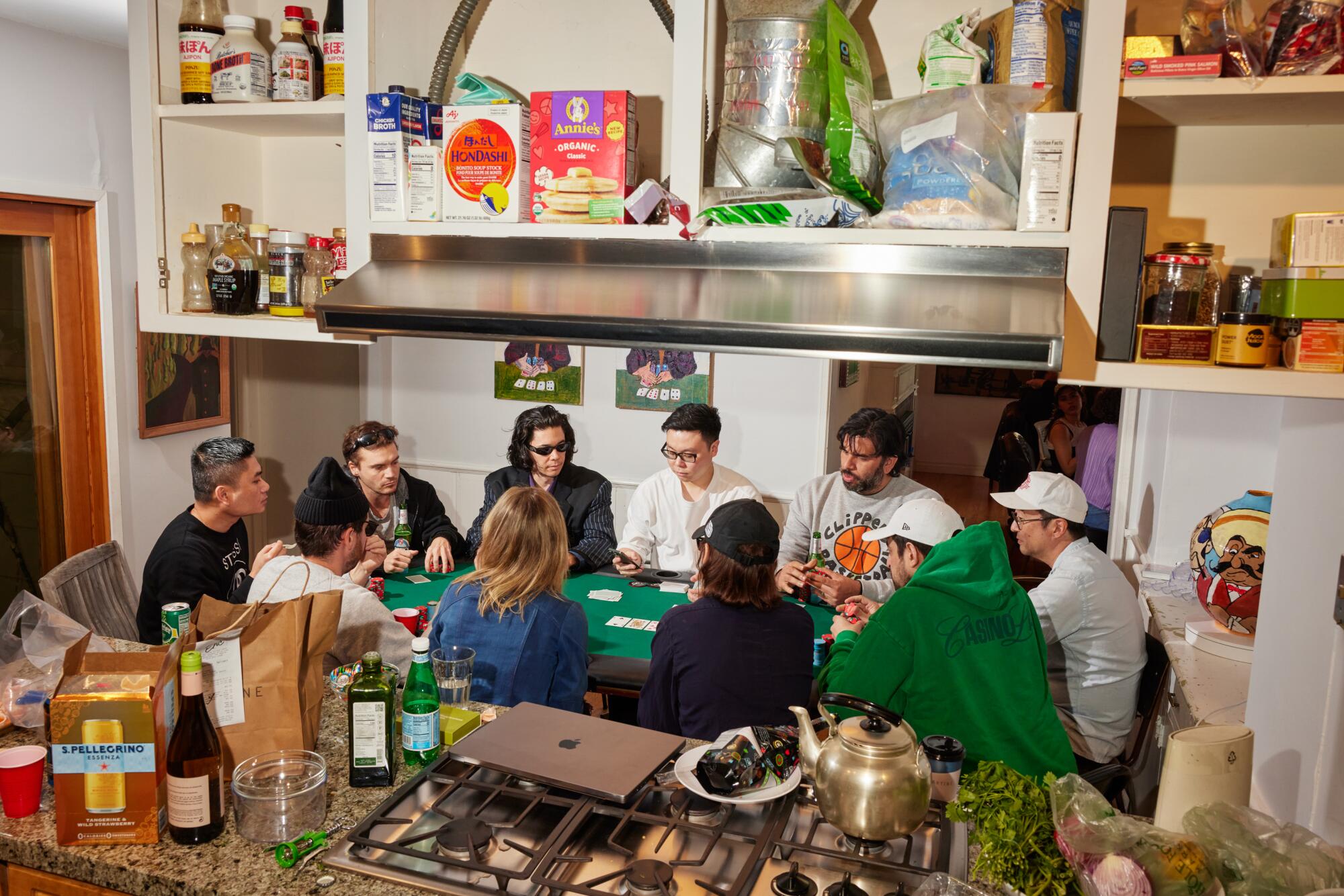 Ten people play poker as seen from the kitchen, with wine, snacks and a laptop on the counter and art above them. 