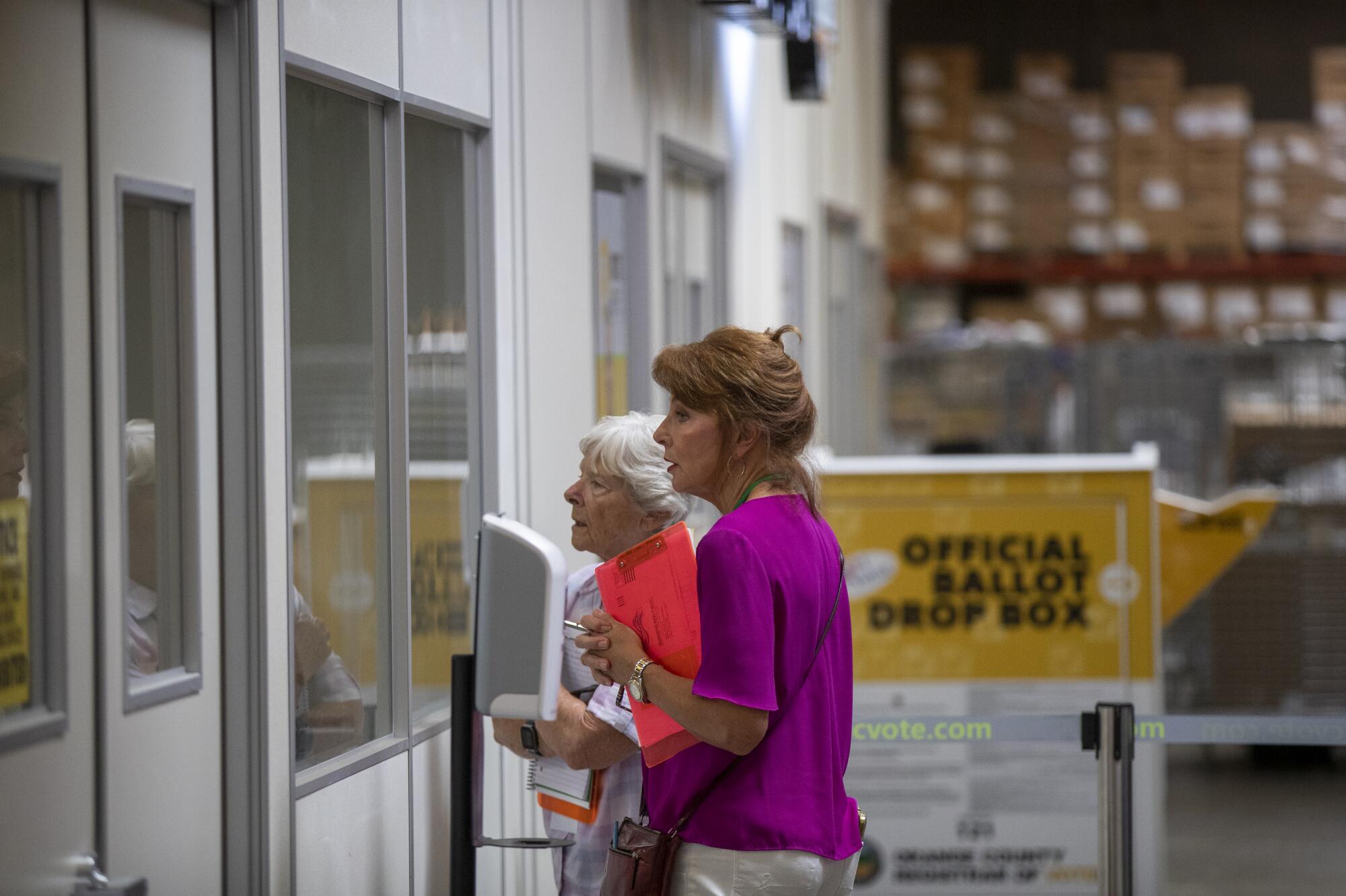 Two women, one in a magenta top and holding an orange clipboard, stand looking through the windows of an office