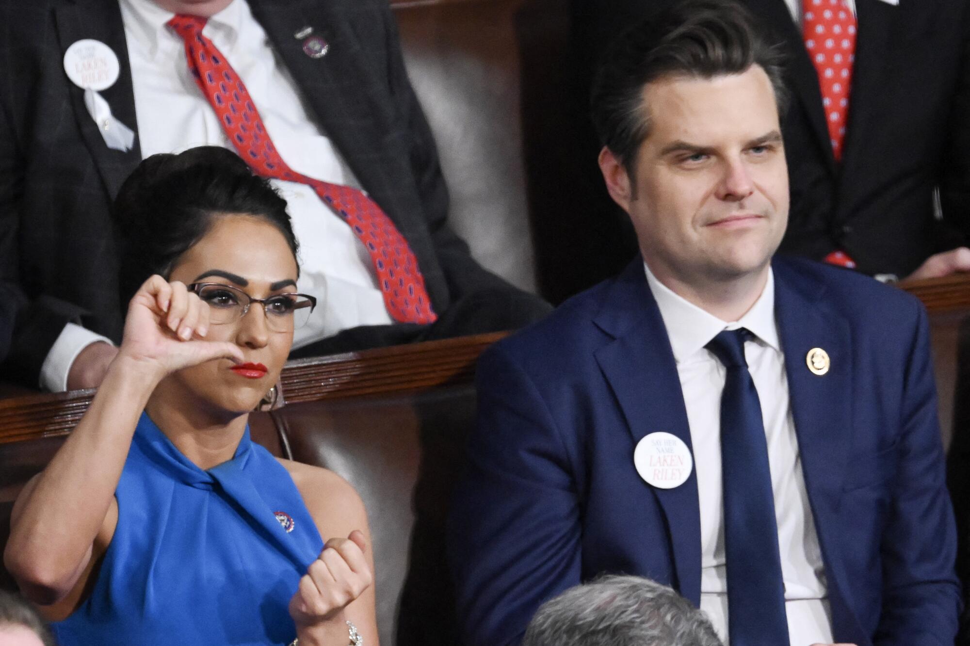 A woman in a blue top and glasses gives a thumbs down next to a man with dark hair, in suit and dark tie
