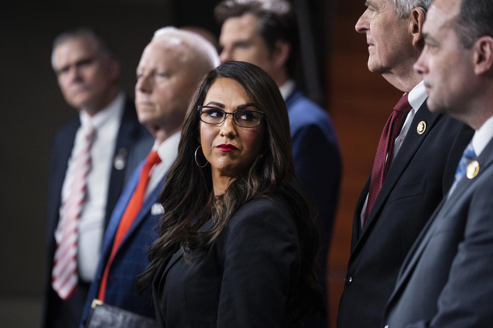 A woman with dark hair, in glasses and dark jacket, is flanked by men in suit and tie 