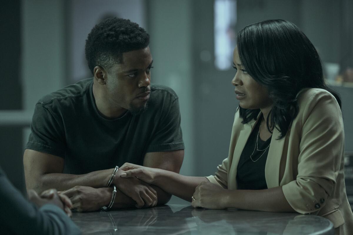 A handcuffed man sitting at a table looks at a woman who has her hand on his.