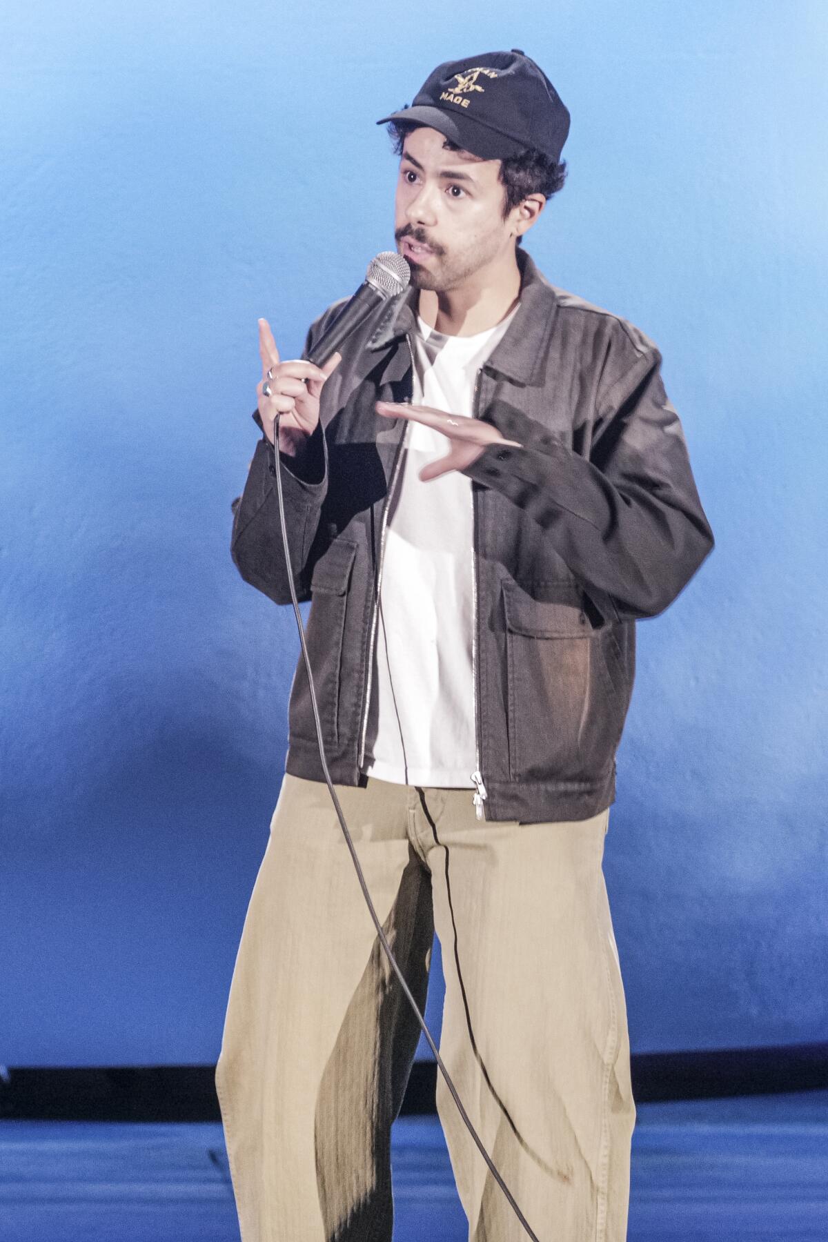 Man in baseball hat and jacket on stage with microphone