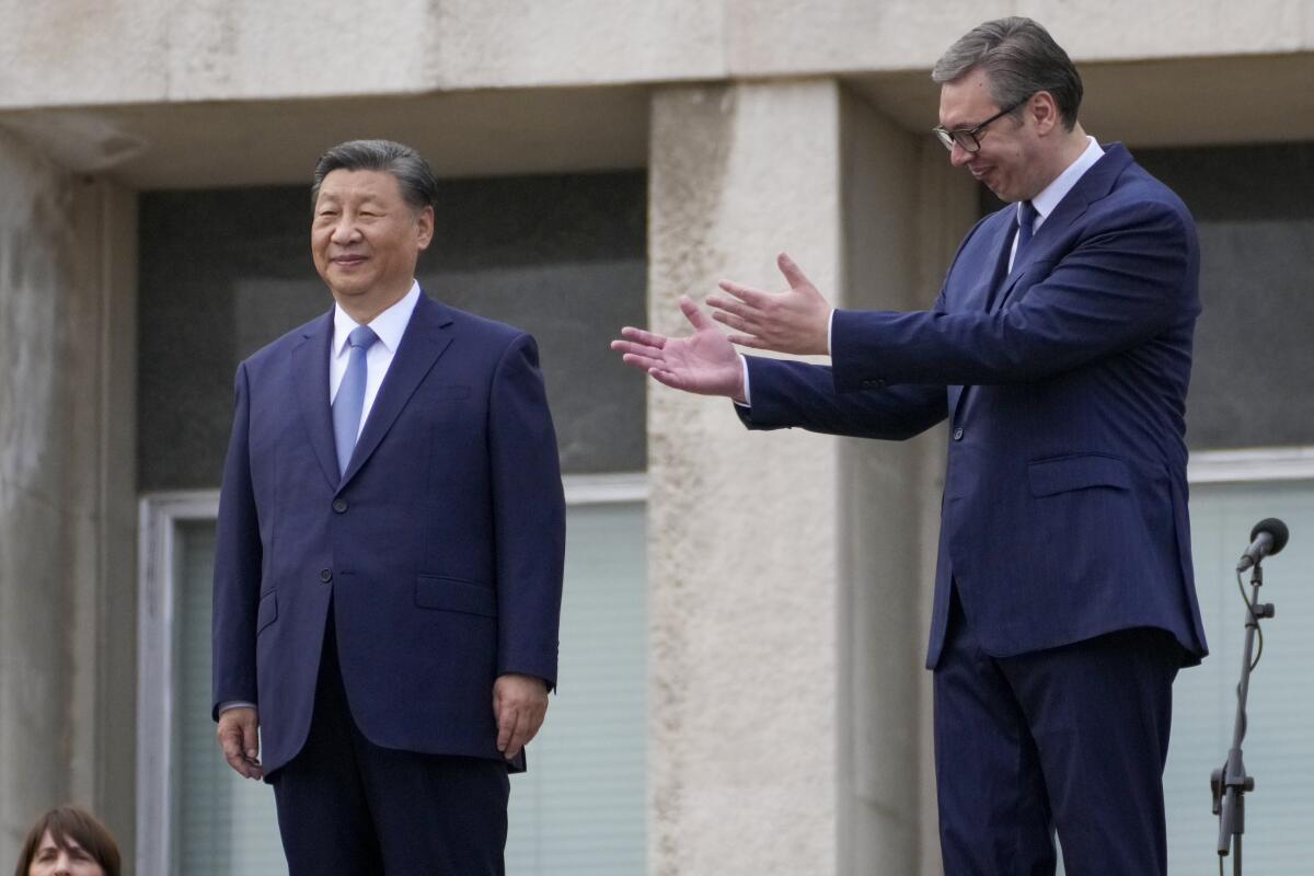 A man in a dark suit and blue tie stands smiling as another man, in dark suit and glasses, holds out his hands toward him
